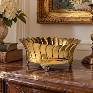 May include: A polished gold-toned brass bowl featuring a scalloped, petal-like rim and a smooth, rounded body. The bowl is supported by three small, ornate feet with intricate detailing, providing a stable base for this decorative metal vessel.