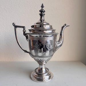 May include: A silver-colored antique tea pot with a lid and a handle. The tea pot has ornate detailing, including a decorative floral pattern and a beaded base. The spout is curved, and the lid is topped with a finial.