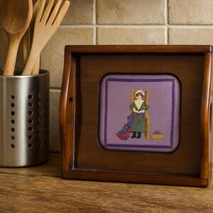 May include: A wooden framed embroidery featuring a woman in a green dress and white bonnet. The embroidery is set against a purple background. A stainless steel utensil holder with wooden spoons and a fork is to the left.