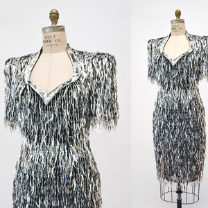 May include: A black and silver sequined dress with fringe detailing. The dress has a sweetheart neckline and short sleeves.