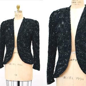 May include: A black sequined jacket with a V-neck and long sleeves. The jacket has a fitted silhouette and a cropped length. The sequins are arranged in a swirling pattern.