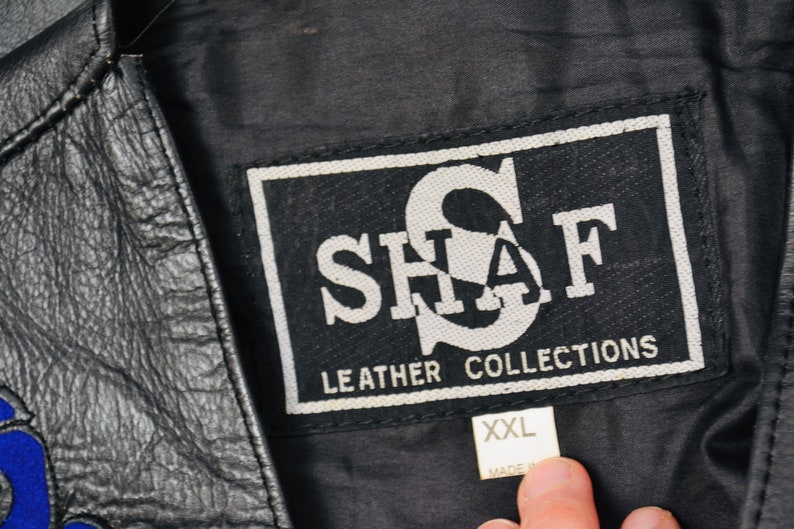 K&ouml;nnte beinhalten: Schwarze Lederjacke mit einem wei&szlig;en Etikett mit der Aufschrift "SHAF LEATHER COLLECTIONS" und einem wei&szlig;en Gr&ouml;&szlig;enetikett mit der Aufschrift "XXL".