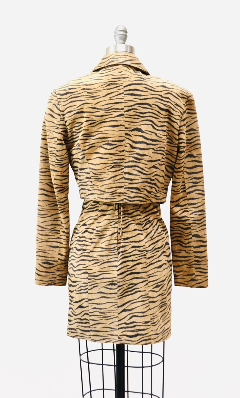 Op de afbeelding: Een tan en zwart tijgerprintjurk met een bijpassend jasje. De jurk heeft een aansluitende pasvorm en een riem in de taille. Het jasje heeft lange mouwen en een kraag.