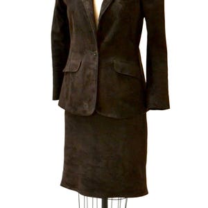 May include: A brown suede blazer and skirt suit on a mannequin. The suit has a single-breasted blazer with a button closure and a skirt that falls to the knee.