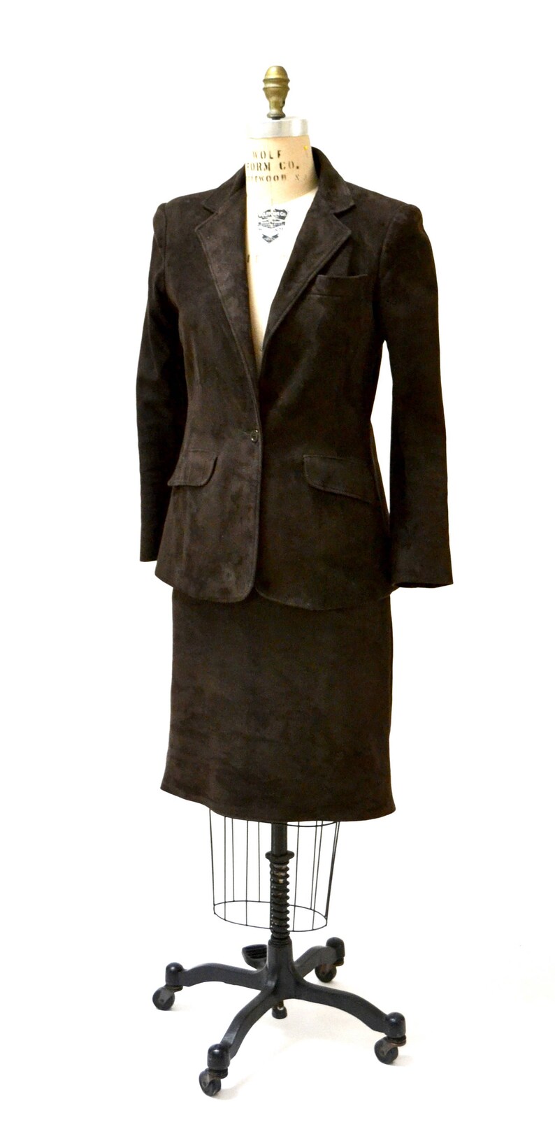 May include: A brown suede blazer and skirt suit on a mannequin. The suit has a single-breasted blazer with a button closure and a skirt that falls to the knee.