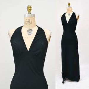 May include: A black halter-neck maxi dress displayed on a vintage dress form. The dress features a deep V-neckline and a fitted bodice that flows into a long, straight skirt. The dress is made of a smooth, solid black fabric.