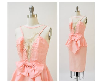May include: A pink dress with a white lace overlay and a large bow at the waist. The dress has a fitted bodice and a full skirt.