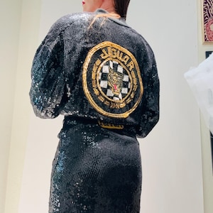 May include: Black sequined jacket and matching skirt. The jacket features a large circular patch on the back with the word "JAGUAR" in gold lettering, a checkered flag design, and a tiger emblem. The outfit is a two-piece set.