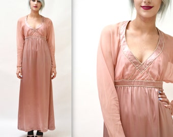 Puede incluir: Un vestido largo, fluido, de color rosa claro con escote en V y mangas largas. El vestido tiene un acabado satinado y está adornado con un ribete de cuentas alrededor del escote y la cintura.