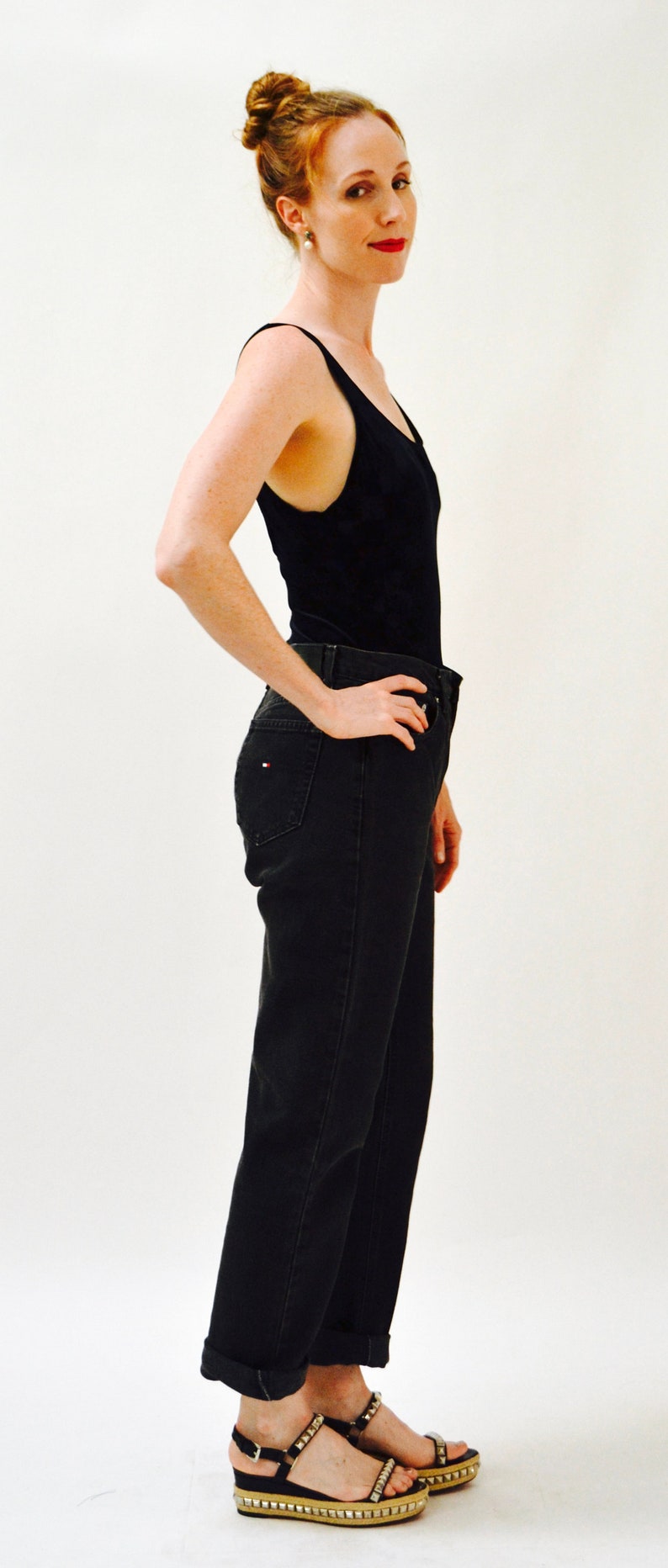 May include: A woman wearing a black tank top and dark wash jeans with a cuffed hem. She is wearing black sandals with silver studs.