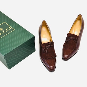 May include: A pair of brown leather loafers with suede fringe detailing and a bow on the top. The shoes are sitting on a green box with the word "GUCCI" printed in gold letters.