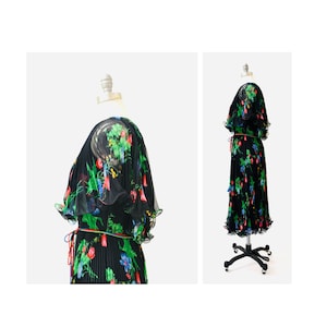May include: A black pleated maxi dress with a floral print in shades of green, red, and blue. The dress has a V-neckline, short sleeves, and a tie at the waist.