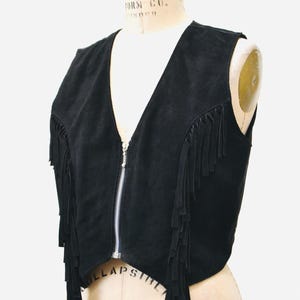 May include: Black suede vest with fringe detailing and a zipper closure. The vest is sleeveless and has a cropped length.