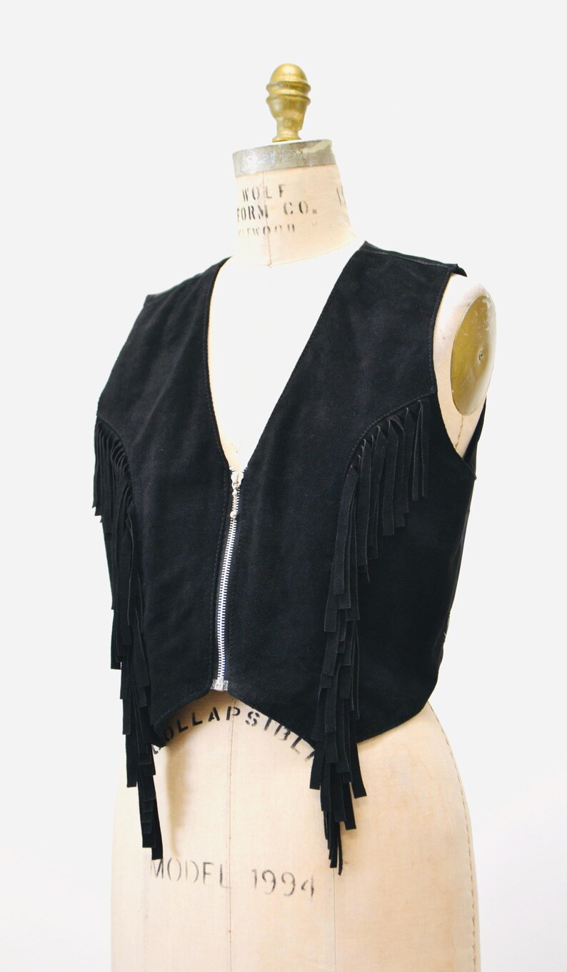May include: Black suede vest with fringe detailing and a zipper closure. The vest is sleeveless and has a cropped length.