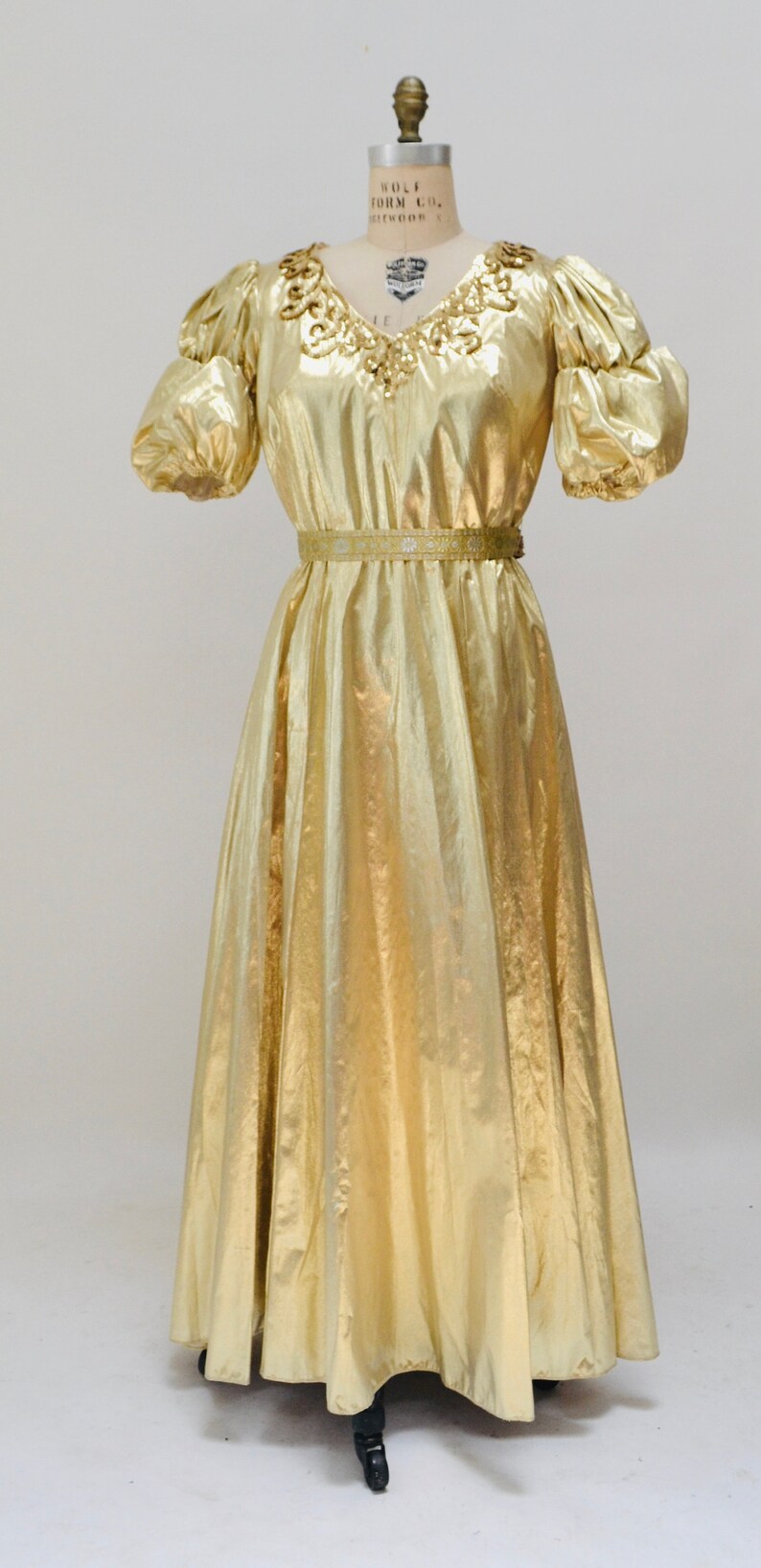 May include: A gold metallic dress with a V-neckline and short puff sleeves. The dress has a beaded embellishment around the neckline and a gold belt.