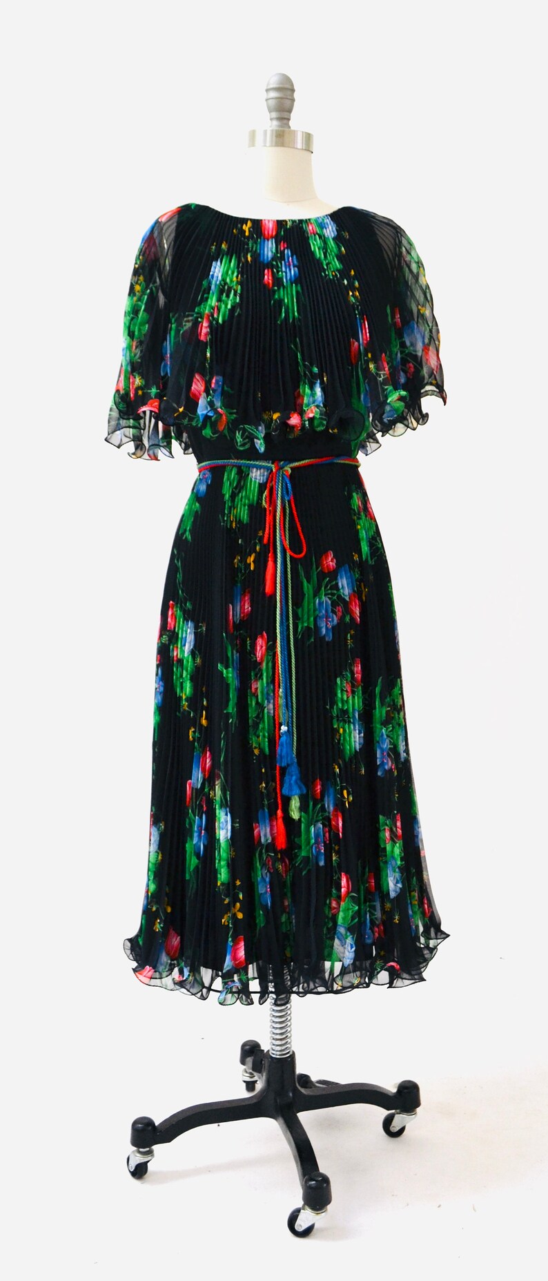 May include: A black pleated dress with a floral print and a colorful tassel tie belt. The dress has short, ruffled sleeves and a flowing skirt.
