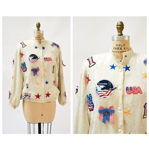 Può includere: Una giacca bomber bianca di paillettes con toppe e decorazioni rosso, bianco e blu. Le toppe includono un segno di pace, stelle, il numero 1 e una toppa che dice "Columbia Eagle-TWA."