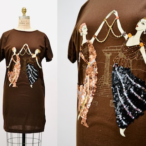 May include: A brown t-shirt with an art deco design. The design features two women in sequined dresses holding a gold chain, set against a gold architectural backdrop. The t-shirt has short sleeves and a round neckline.