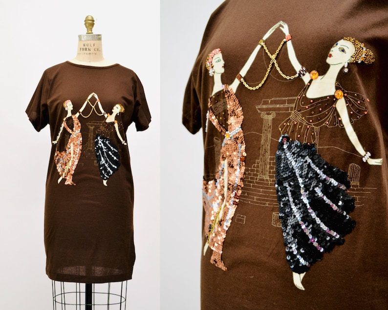 May include: A brown t-shirt with an art deco design. The design features two women in sequined dresses holding a gold chain, set against a gold architectural backdrop. The t-shirt has short sleeves and a round neckline.
