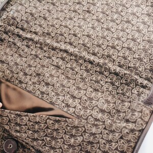 May include: The inside of a brown leather bag with a butterfly pattern lining. The lining is a light brown color with a darker brown butterfly pattern.