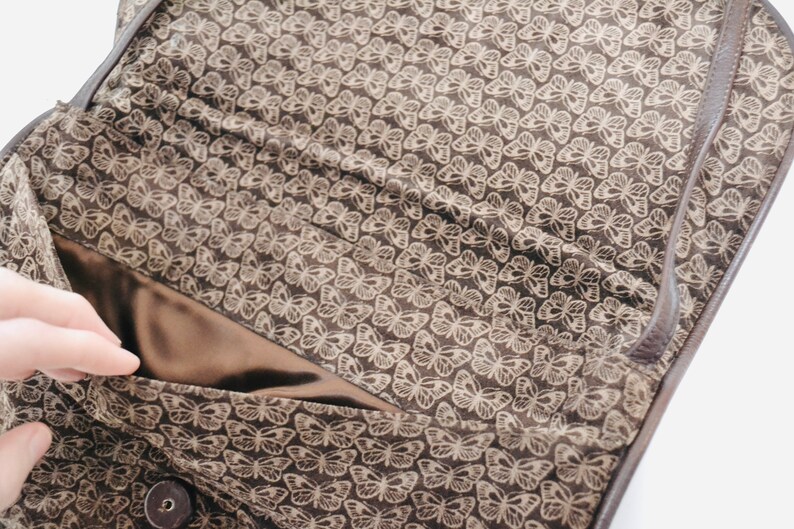 May include: The inside of a brown leather bag with a butterfly pattern lining. The lining is a light brown color with a darker brown butterfly pattern.