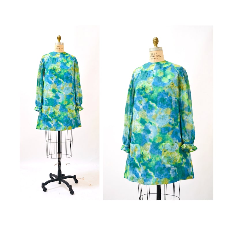 May include: A vintage 1960s mod dress with a blue, green, and yellow abstract print. The dress has long sleeves with ruffles at the cuffs and a round neckline. The dress is made of a lightweight fabric and has a loose fit.