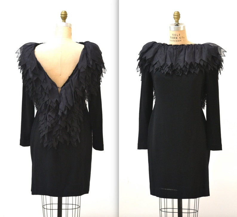 May include: A black dress with a ruffled neckline and a deep V-back. The ruffles are made of a sheer fabric and are layered to create a dramatic effect.