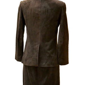 May include: A brown suede suit jacket and skirt set. The jacket has a notched collar and long sleeves. The skirt is knee-length and has a straight silhouette.