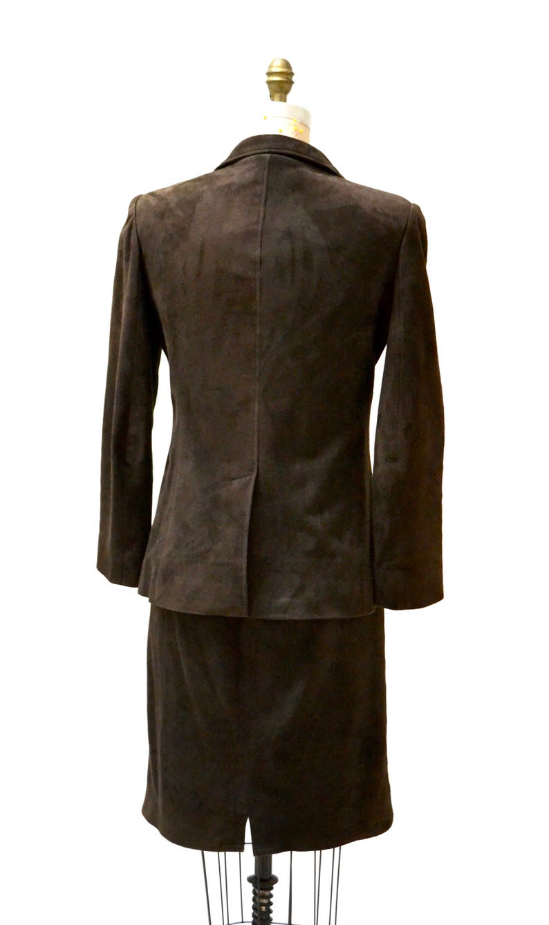 May include: A brown suede suit jacket and skirt set. The jacket has a notched collar and long sleeves. The skirt is knee-length and has a straight silhouette.