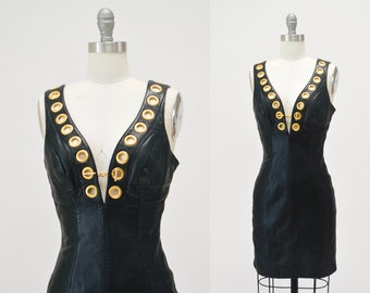 May include: A black leather mini dress with a deep V-neckline and gold grommets. The dress has a sleeveless design and a fitted silhouette.