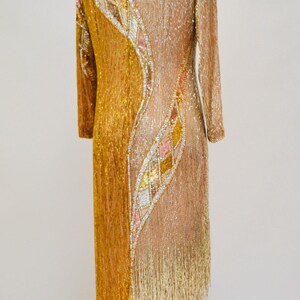 Op de afbeelding: Een gouden en beige parelkleed met een geometrisch patroon en franjes. De jurk is op een mannequin en heeft lange mouwen.