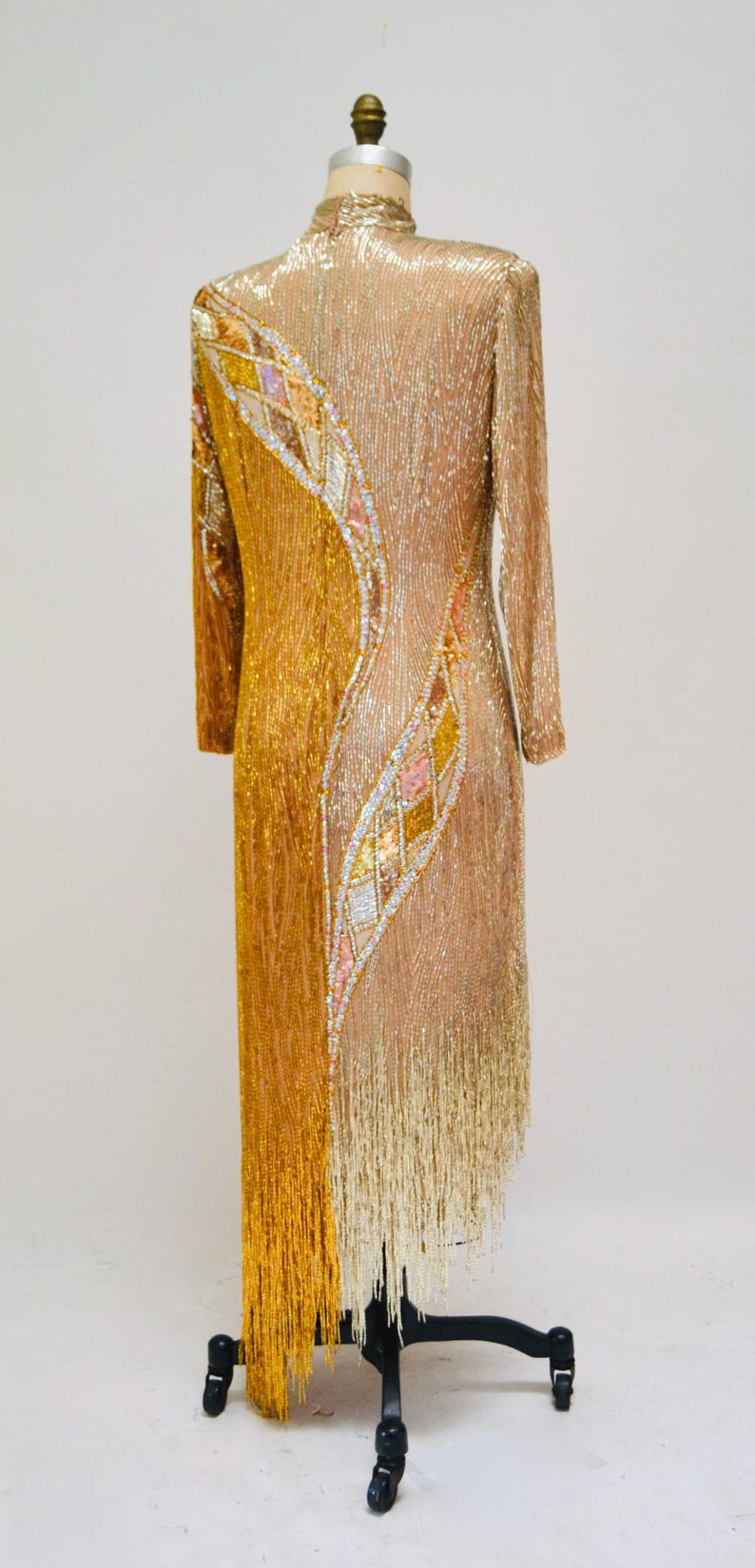 Op de afbeelding: Een gouden en beige parelkleed met een geometrisch patroon en franjes. De jurk is op een mannequin en heeft lange mouwen.