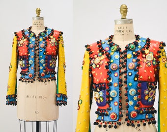 May include: A colourful, multi-coloured, beaded and embroidered jacket with a yellow collar and sleeves. The jacket has a red, blue and purple patchwork design with a variety of embellishments, including beads, sequins and pom-poms.