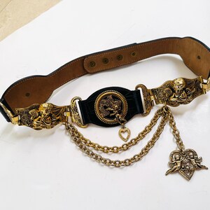 Puede incluir: Un cinturón de cuero negro con herrajes metálicos dorados y motivos de querubines. La pieza central es un medallón circular con un dije de corazón, conectado a dos cadenas doradas drapeadas y un colgante en forma de corazón con querubines.