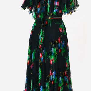 May include: A black pleated dress with a floral print featuring red, blue, and green flowers. The dress has a short, cape-like top and a skirt that falls to the knees. The dress is on a mannequin with a black stand.