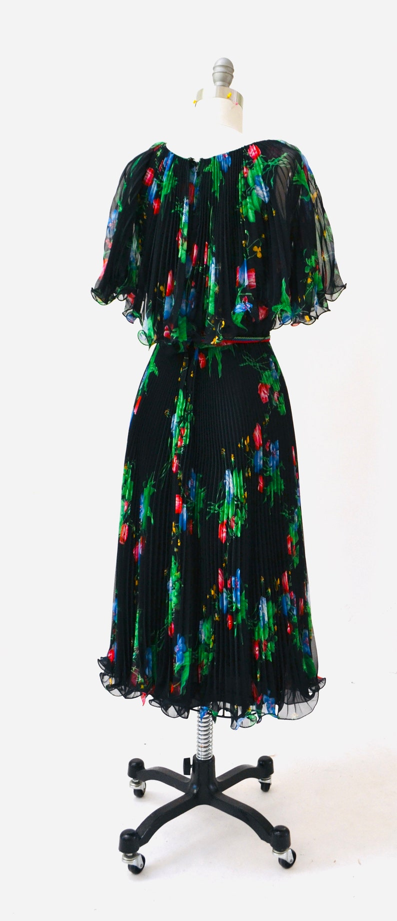 May include: A black pleated dress with a floral print featuring red, blue, and green flowers. The dress has a short, cape-like top and a skirt that falls to the knees. The dress is on a mannequin with a black stand.