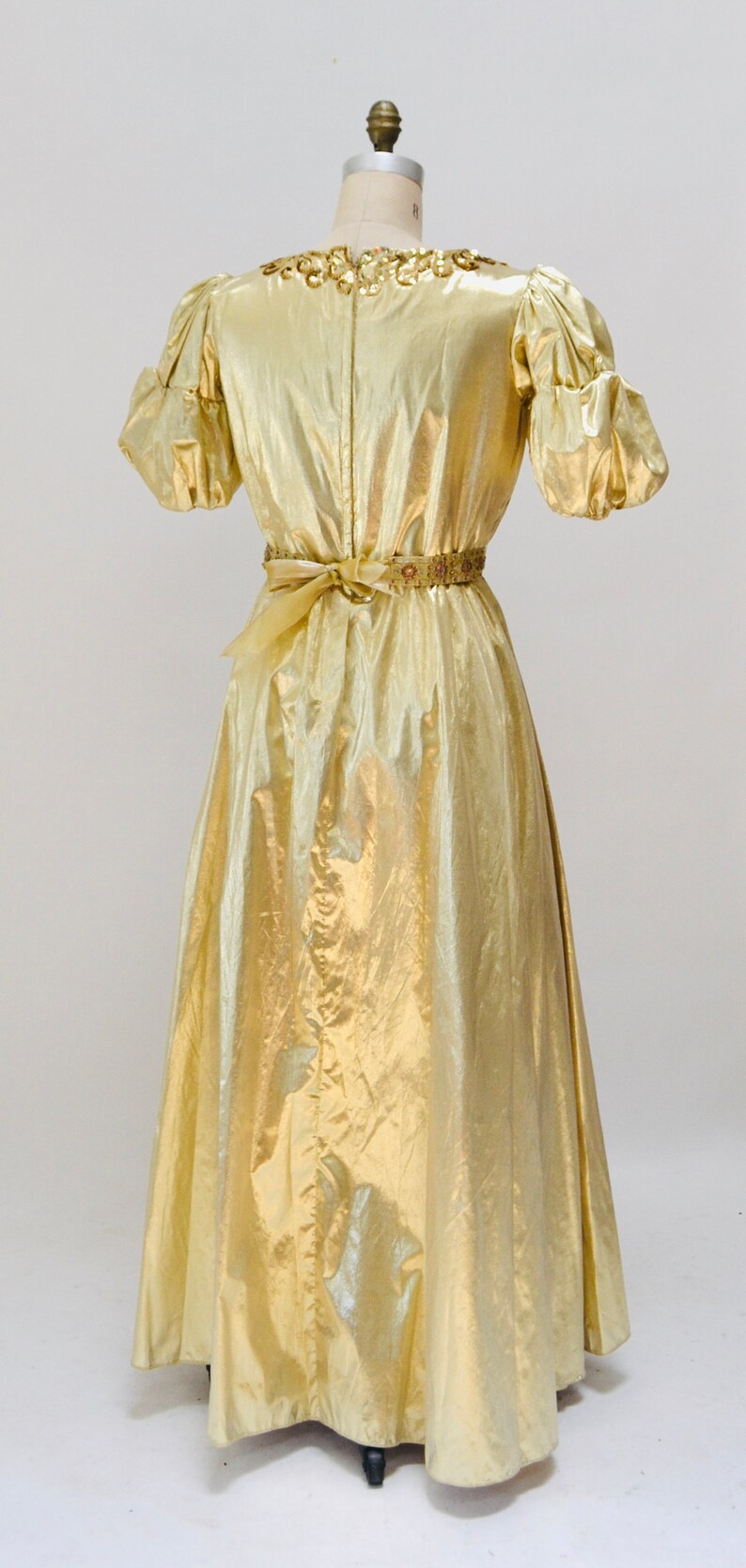 May include: A gold metallic dress with a gathered neckline and short puff sleeves. The dress has a gold beaded belt and a flowing skirt.