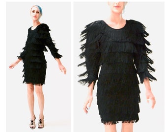 May include: A black dress with fringe detailing on the sleeves and skirt. The dress is knee-length and has a scoop neckline.