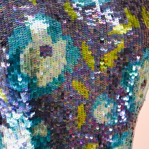 May include: Close-up of a fabric with a blue, green, and purple iridescent sequin pattern. The sequins are arranged in a floral design.