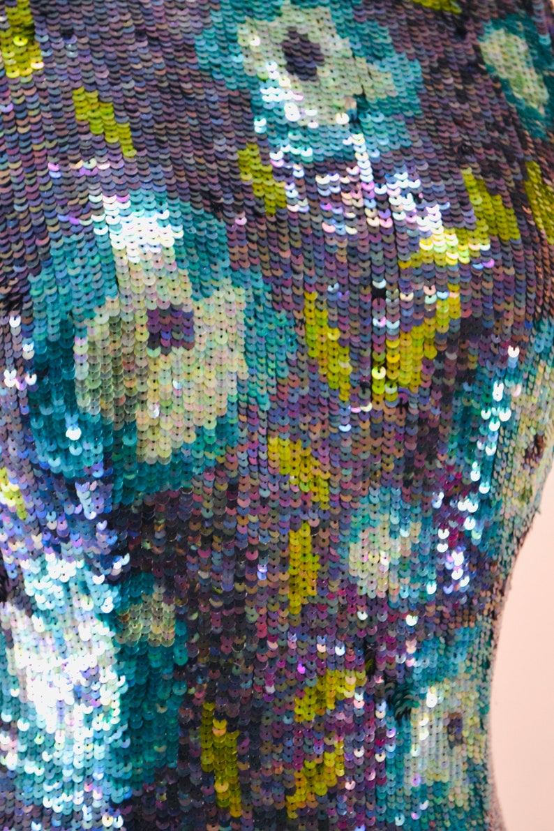 May include: Close-up of a fabric with a blue, green, and purple iridescent sequin pattern. The sequins are arranged in a floral design.
