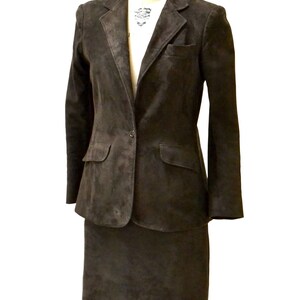 May include: A brown suede suit jacket and skirt set. The jacket has a single button closure and two pockets. The skirt is knee-length.