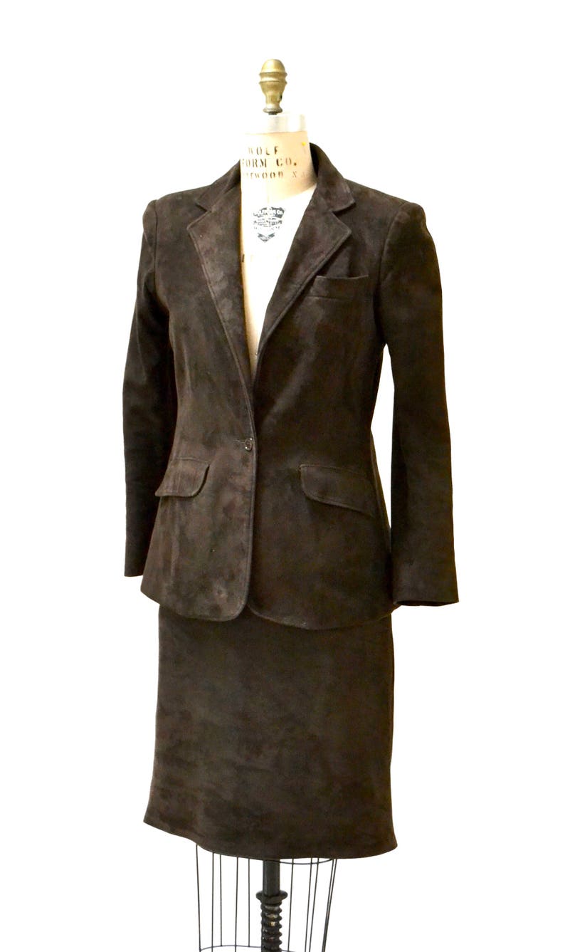 May include: A brown suede suit jacket and skirt set. The jacket has a single button closure and two pockets. The skirt is knee-length.