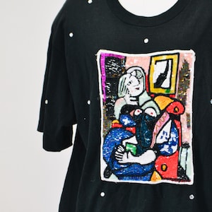 May include: Black t-shirt with a colorful embroidered image of two figures in a room. The image is surrounded by a white border and the shirt is decorated with small white dots.