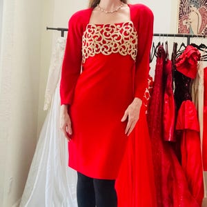 May include: A red dress with a gold embroidered bodice and matching red bolero jacket. The dress is knee-length and has long sleeves. The bolero has long sleeves and a square neckline. The outfit is paired with black tights.