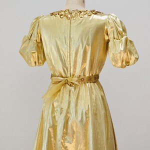 May include: A gold metallic dress with a beaded neckline and a matching beaded belt. The dress has a fitted bodice and a flowing skirt.