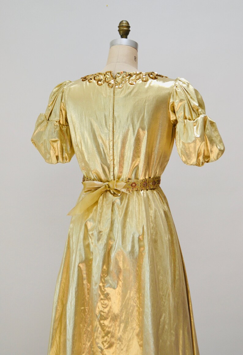 May include: A gold metallic dress with a beaded neckline and a matching beaded belt. The dress has a fitted bodice and a flowing skirt.