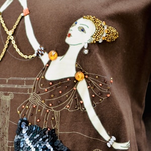 May include: A brown t-shirt with an Art Deco design of a woman in a gown. The illustration has gold and orange bead details, a sequined skirt, and a gold chain. The woman's hair is decorated with gold beads.