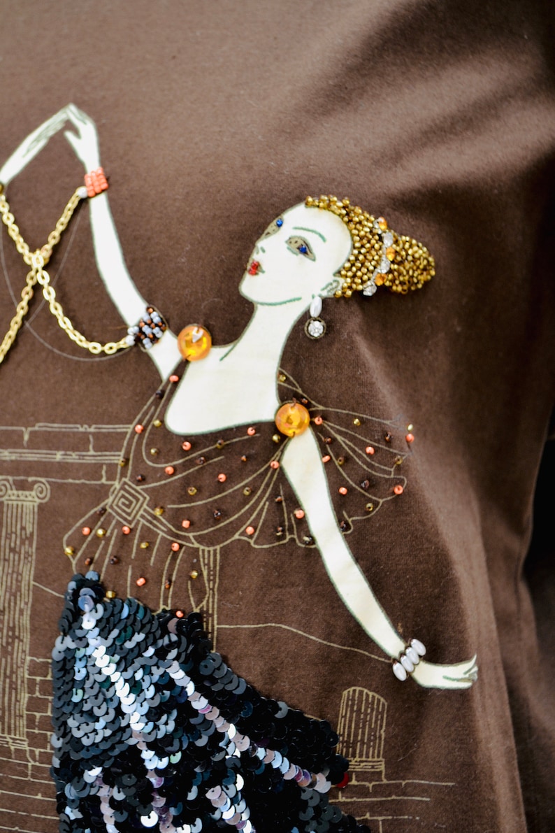 May include: A brown t-shirt with an Art Deco design of a woman in a gown. The illustration has gold and orange bead details, a sequined skirt, and a gold chain. The woman's hair is decorated with gold beads.