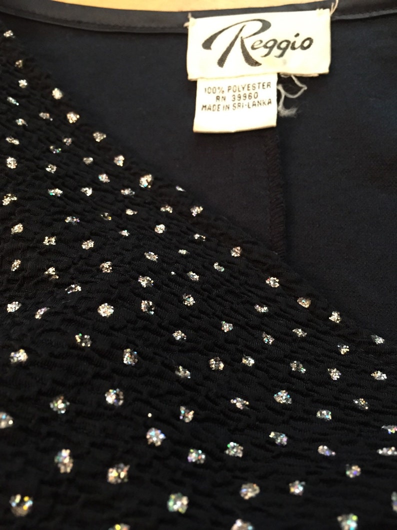 May include: A black top with a V-neckline and a sparkly, textured fabric. The top has a white label with the text "Reggio" and "100% Polyester RN 39960 Made in Sri Lanka".