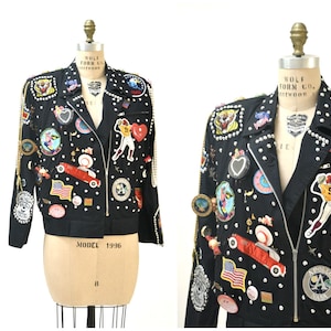 Op de afbeelding: Een zwart leren jack met een ritssluiting en talloze patches en versieringen. Het jack heeft modelnummer 1996.